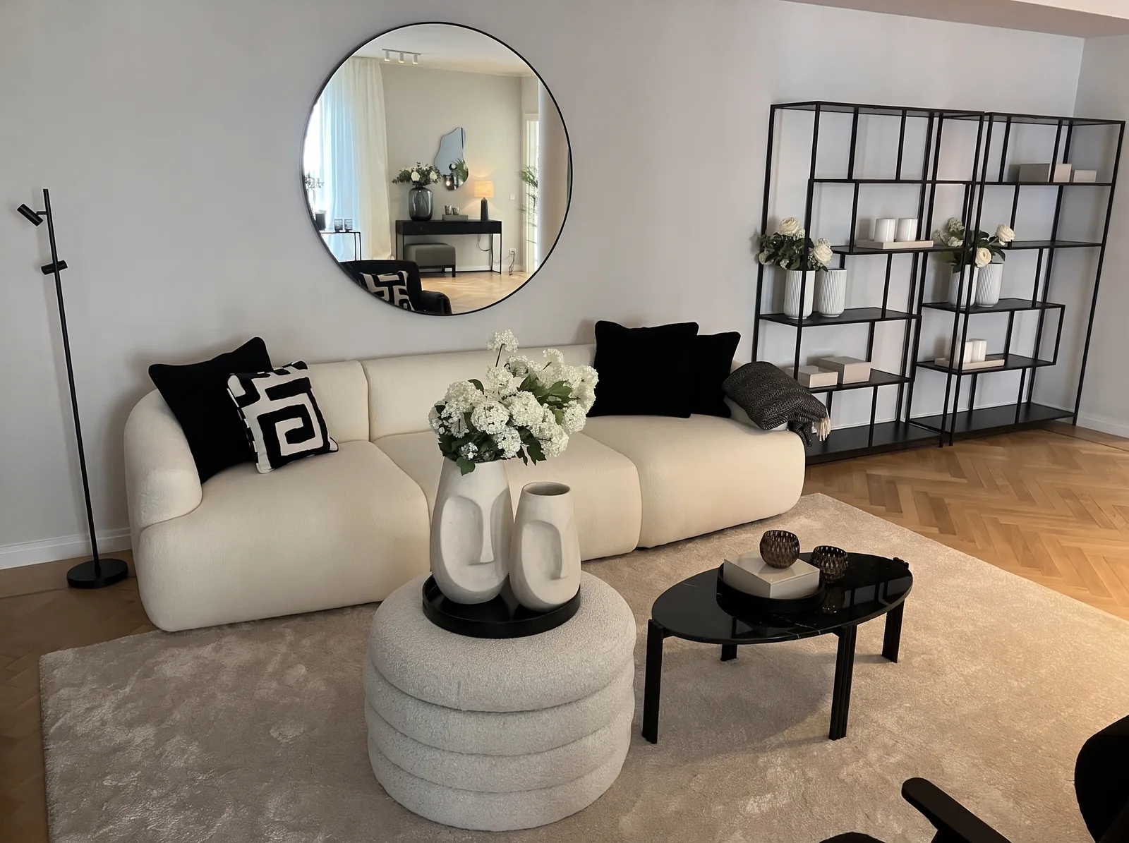 Eingerichtetes Wohnzimmer: cremefarbenes Sofa, runder Spiegel, schwarzes Regal mit Blumen, Couchtisch aus schwarzem Marmor