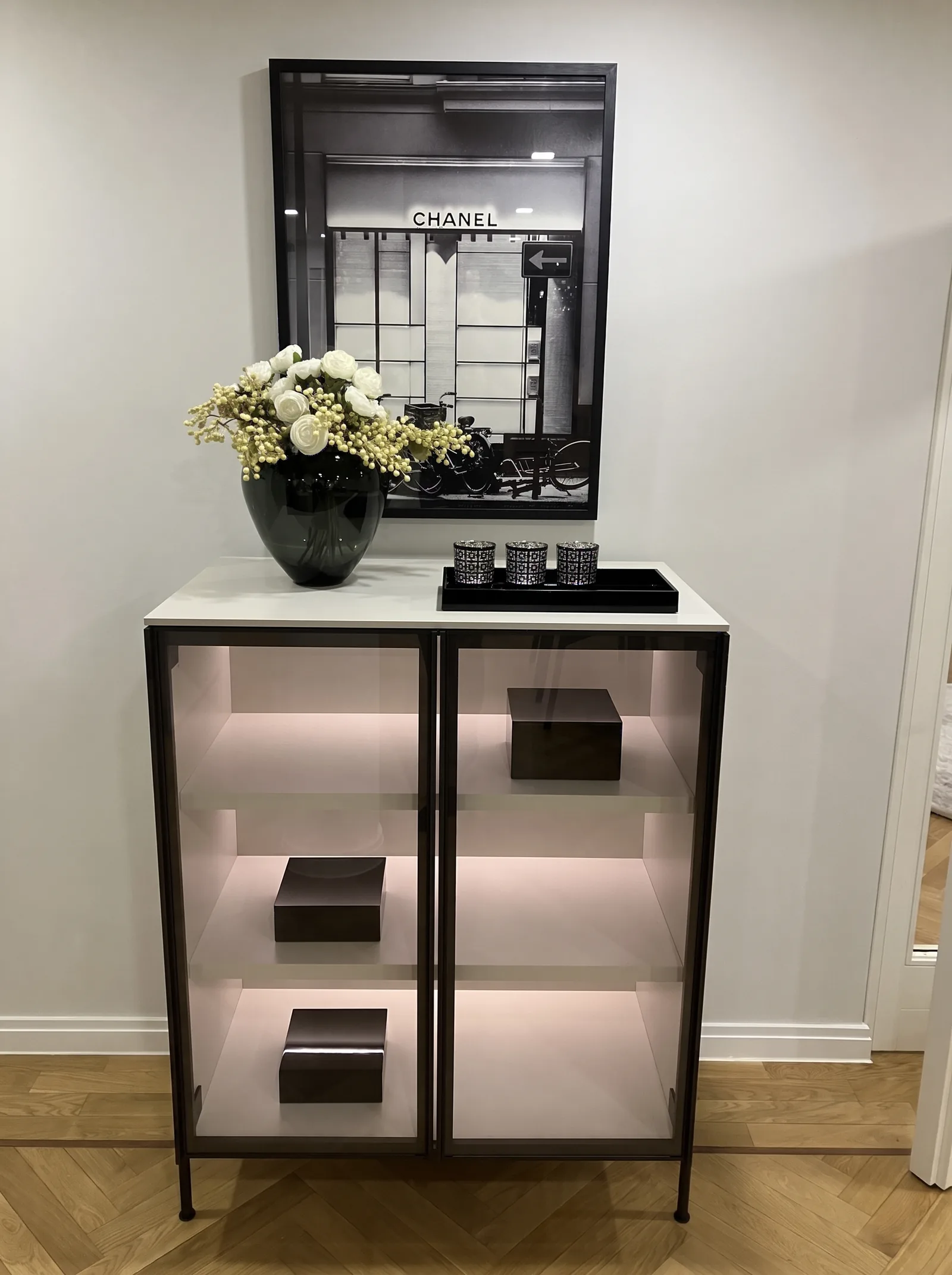 Vitrine mit Glastüren und beleuchteten Fachböden, darüber Blumen und ein Schwarz-Weiß-Bild