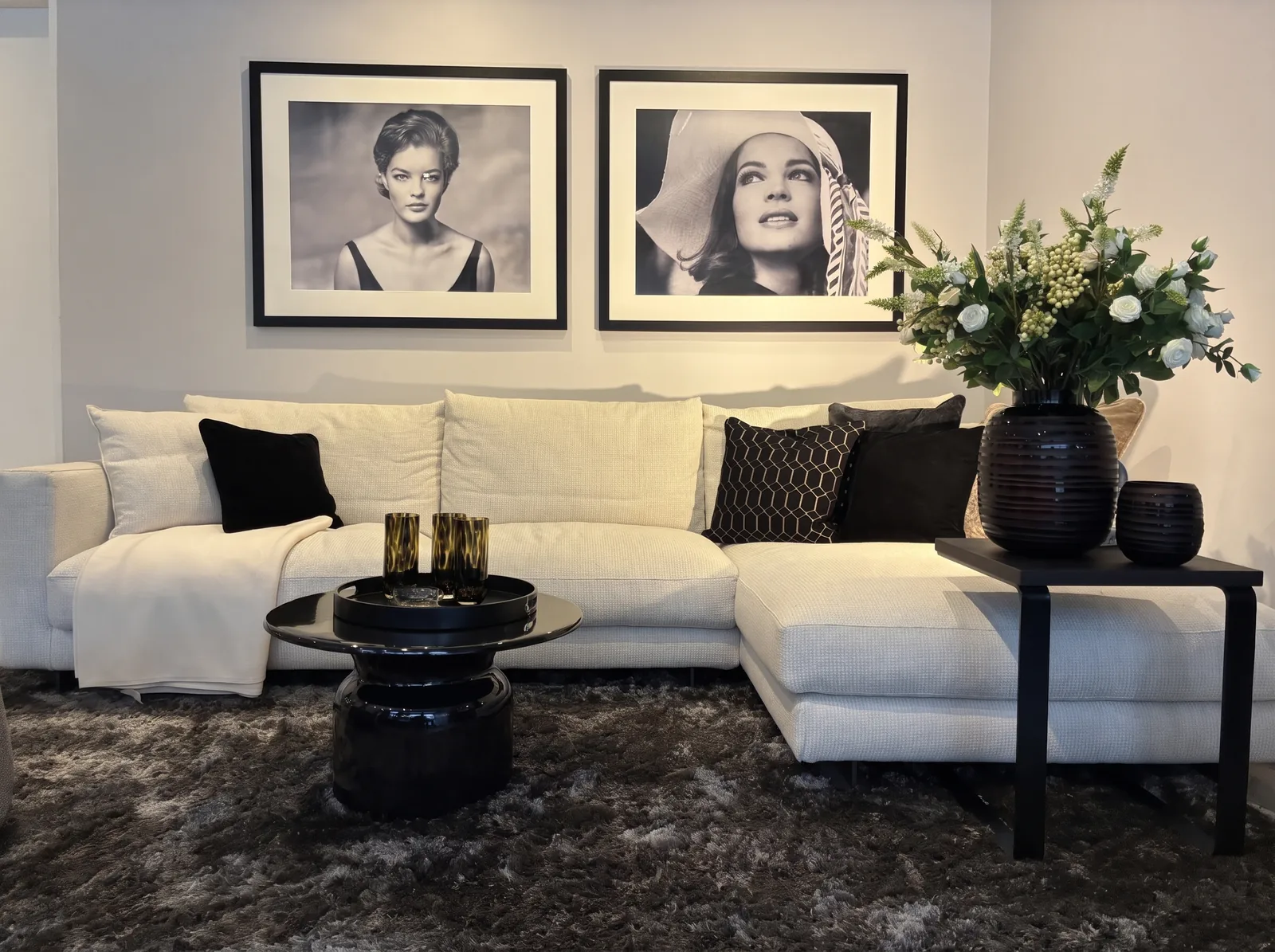Wohnzimmer im Showroom: helles Ecksofa, schwarzer Couchtisch, zwei gerahmte Schwarz-Weiß-Porträts